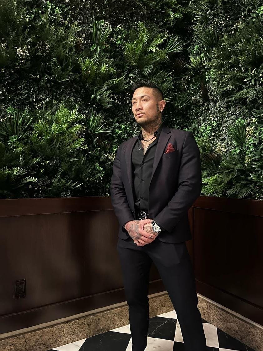 Eros in a tailored black suit against a green wall